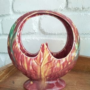 Casey Ware basket vase in wine red with yellow and green glaze, hoop handle and leaf-moulded sides, against a white brick wall
