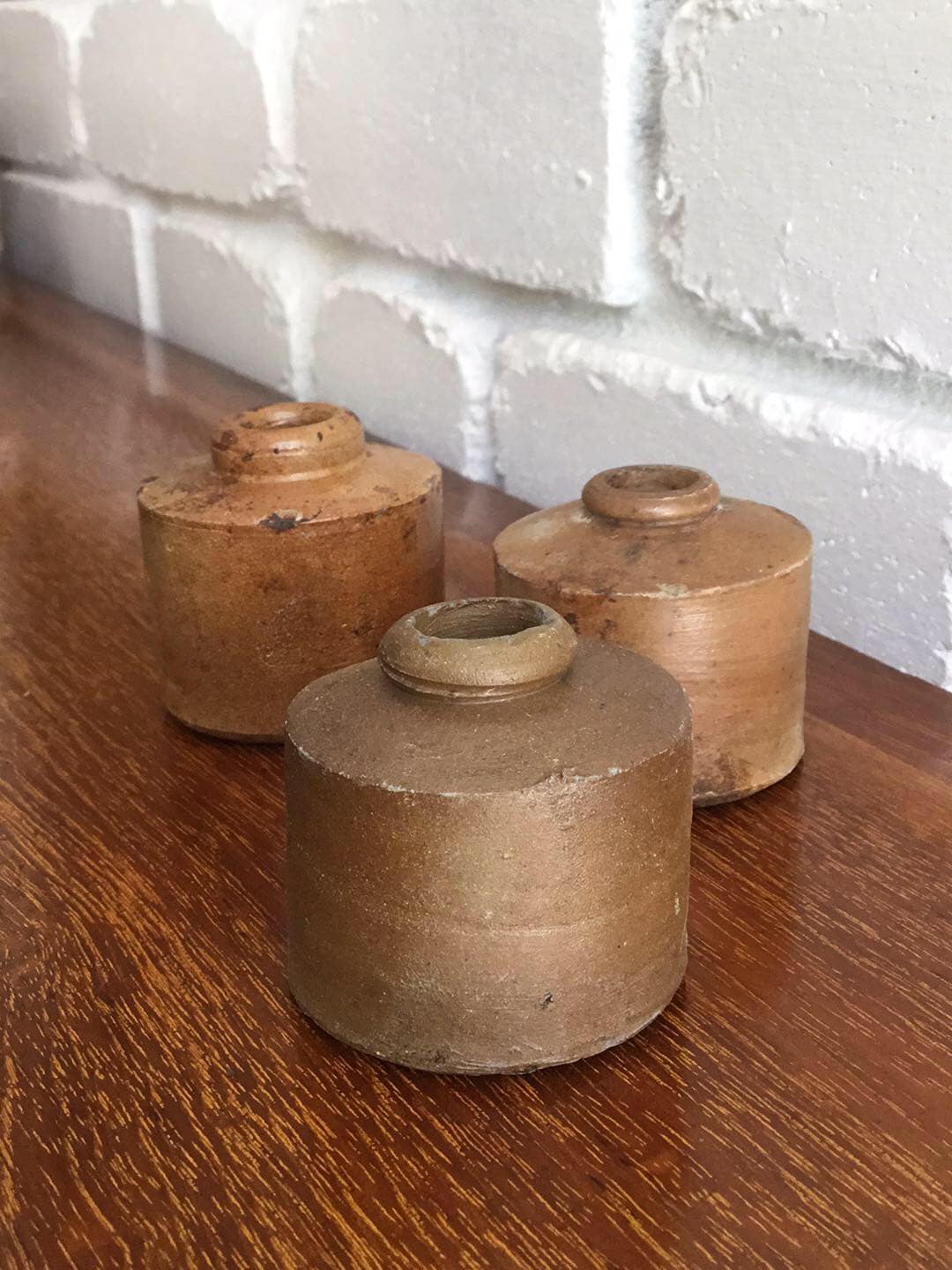 Set of three antique salt glazed stoneware penny inkwells in honey and tan browns on a timber sideboard against a white brick wall