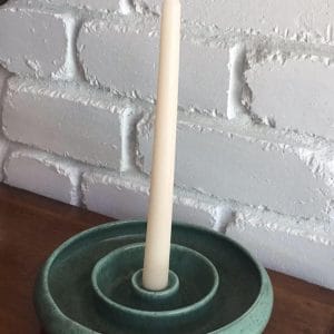 Guy Boyd speckled green candlestick holder with a cream taper candle on a timber sideboard against a white brick wall